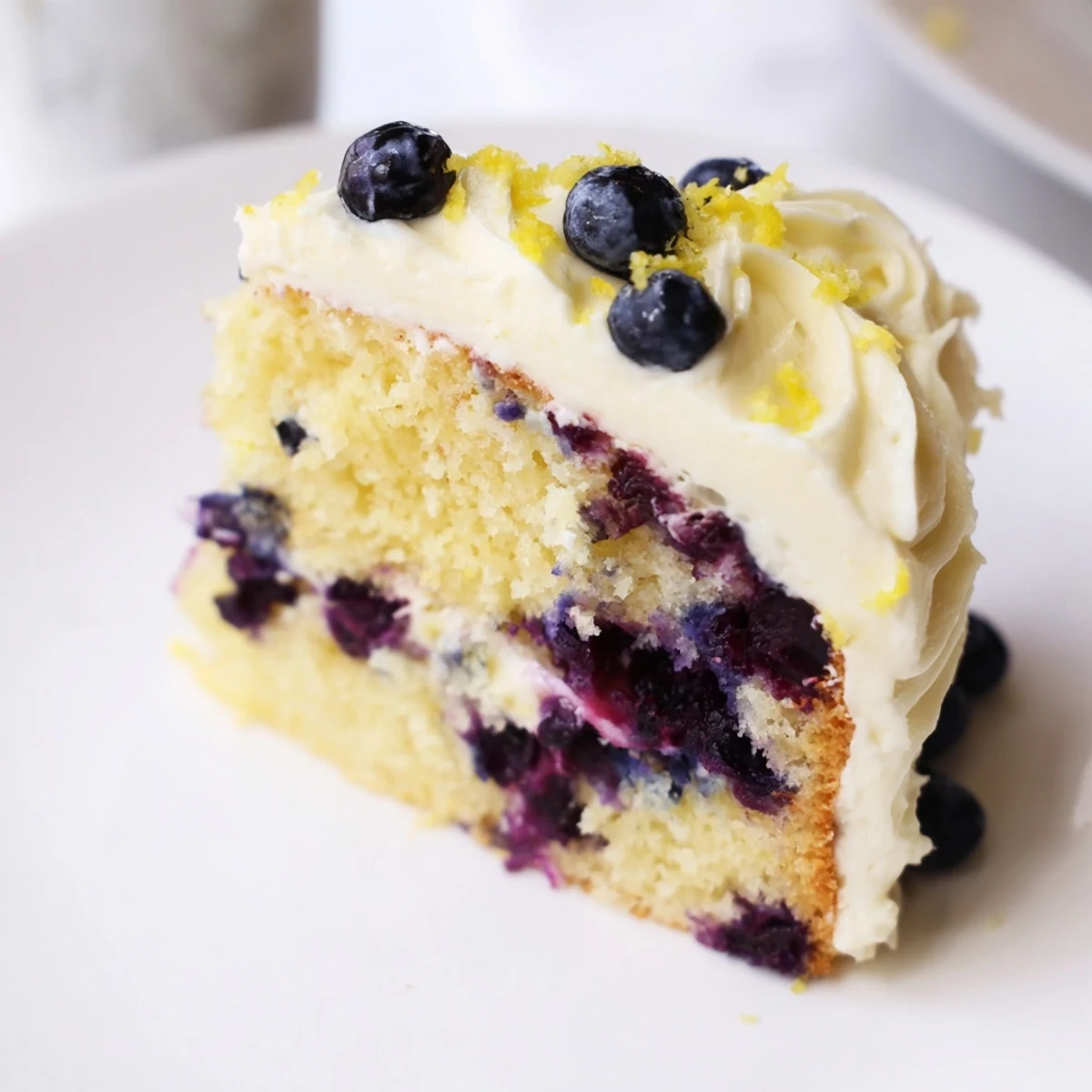 A slice of homemade Lemon Blueberry Cake with frosting is served on a white plate, revealing a moist crumb studded with fresh berries.