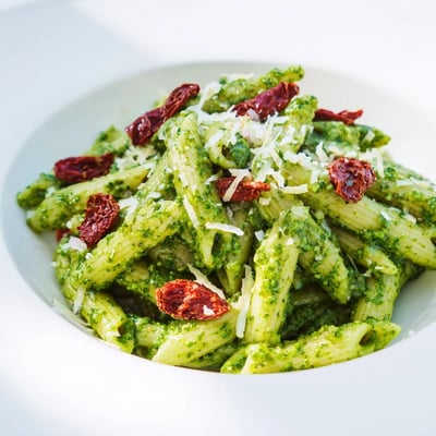 A close-up of Green Pesto Pasta with Sun-Dried Tomatoes, glistening with olive oil and fresh Parmesan garnish.