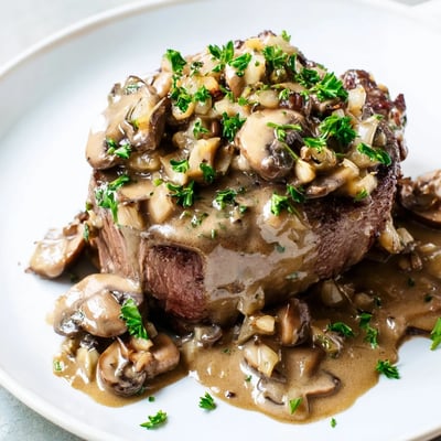 A fork tenderly cuts into beef tenderloin, revealing juicy pink interior beneath the rich, velvety brandy-infused mushroom sauce.  